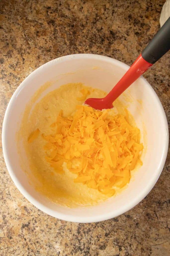 A bowl with a mixture and shredded cheddar cheese being stirred with a red silicone spatula.