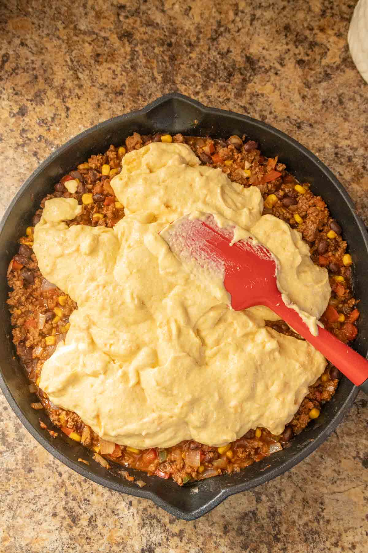 Spreading cornbread batter over a meat and vegetable mixture in a cast-iron skillet.