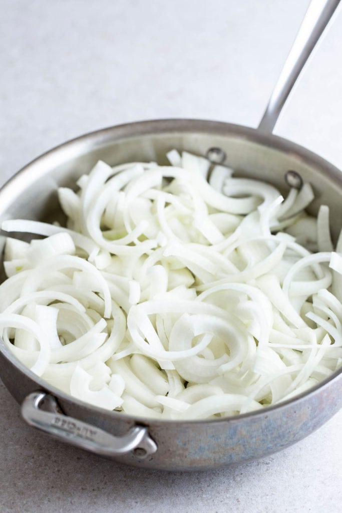 Sliced onions in a stainless steel frying pan on a kitchen countertop.