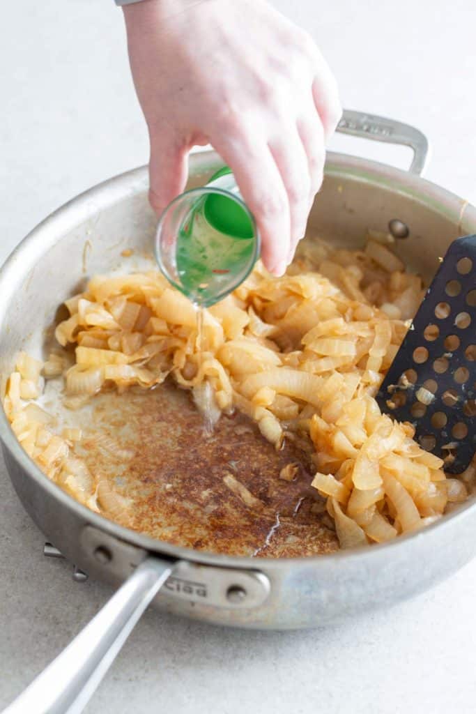 A person's hand pouring liquid into a pan of sautéed onions.