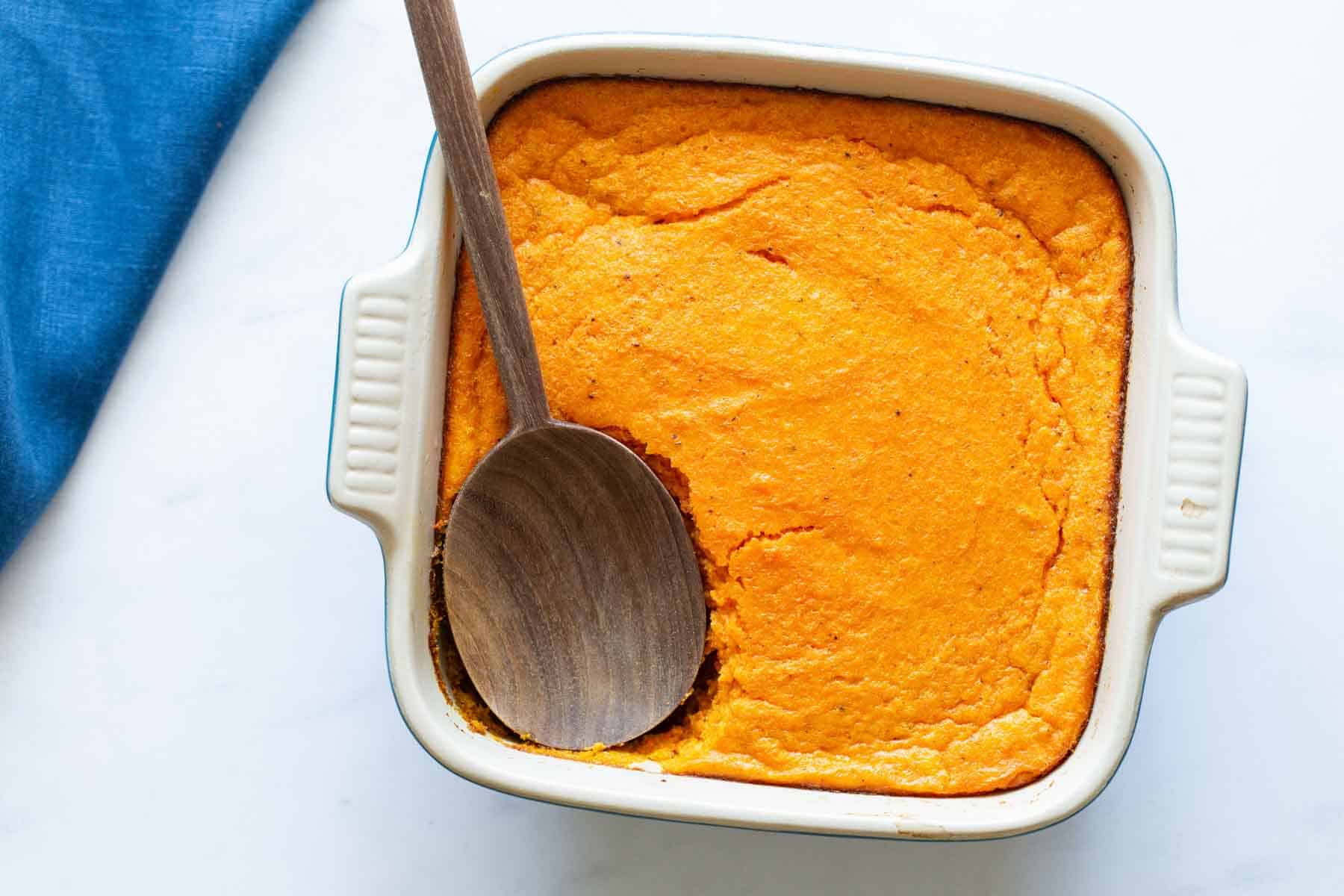 Freshly baked carrot souffle in a square dish with a wooden spoon.