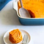 A serving of casserole on a white plate with a fork, next to a baking dish with more casserole and a wooden serving spoon.