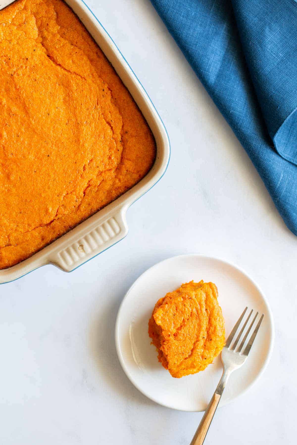 A serving of carrot souffle on a white plate with a fork, beside a full casserole dish on a kitchen counter.