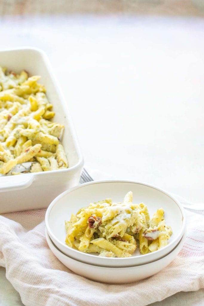 A plate of creamy penne pasta topped with cheese and herbs, beside a larger dish of the same pasta, set on a light-colored tablecloth.