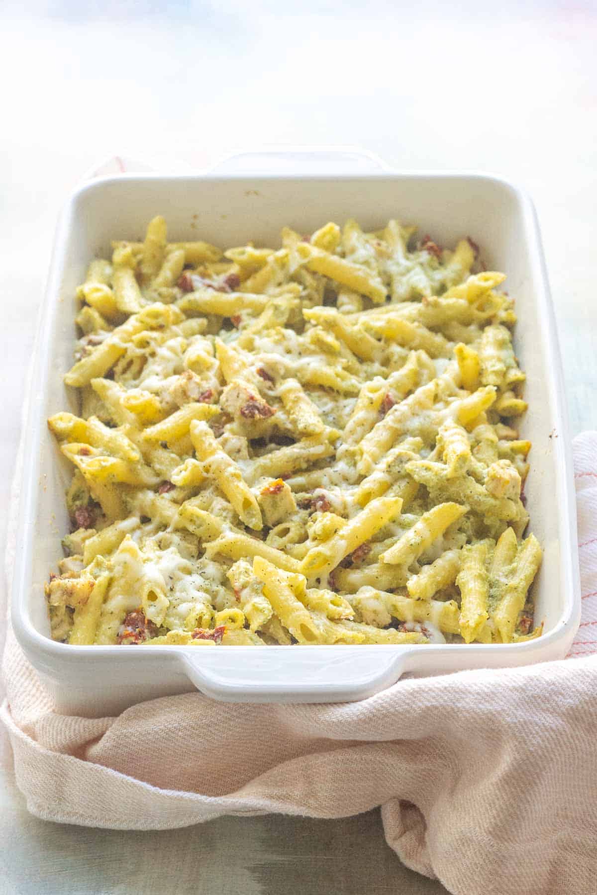 A baked pasta dish with melted cheese and herbs in a white rectangular casserole dish, resting on a beige cloth.