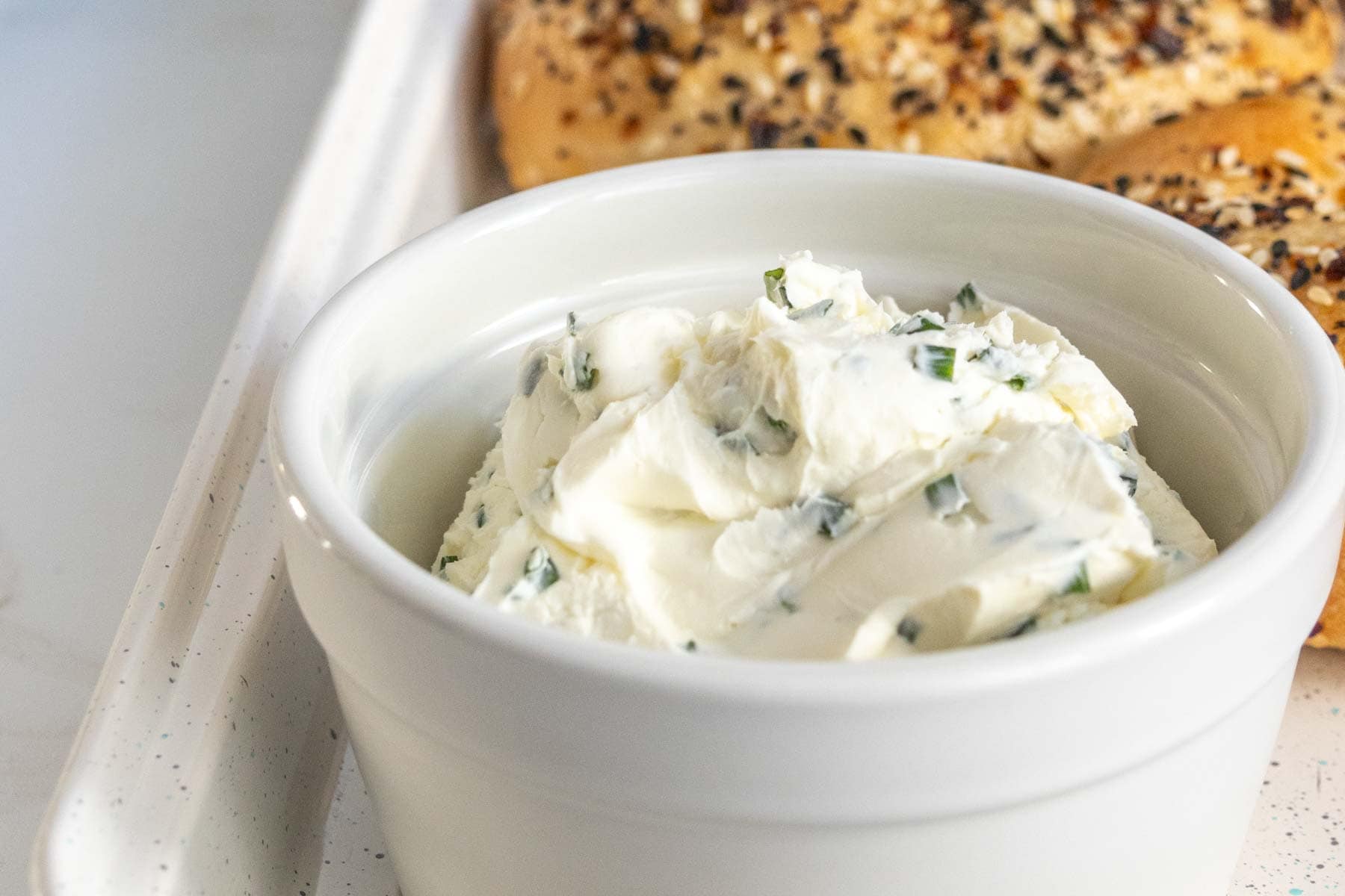 A white bowl of herb-infused cream cheese beside a bagel on a light-colored countertop.