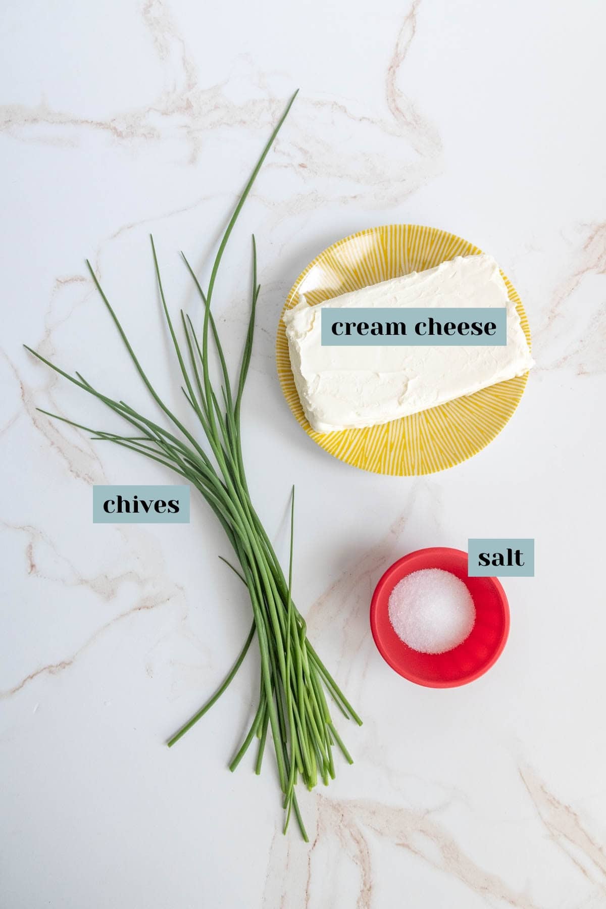 A container of cream cheese, fresh chives, and a small red bowl of salt on a marble surface.