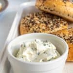 A bowl of herb cream cheese with a tray of everything bagels in the background, served on a kitchen counter.