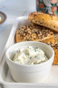 A bowl of herb cream cheese with a tray of everything bagels in the background, served on a kitchen counter.