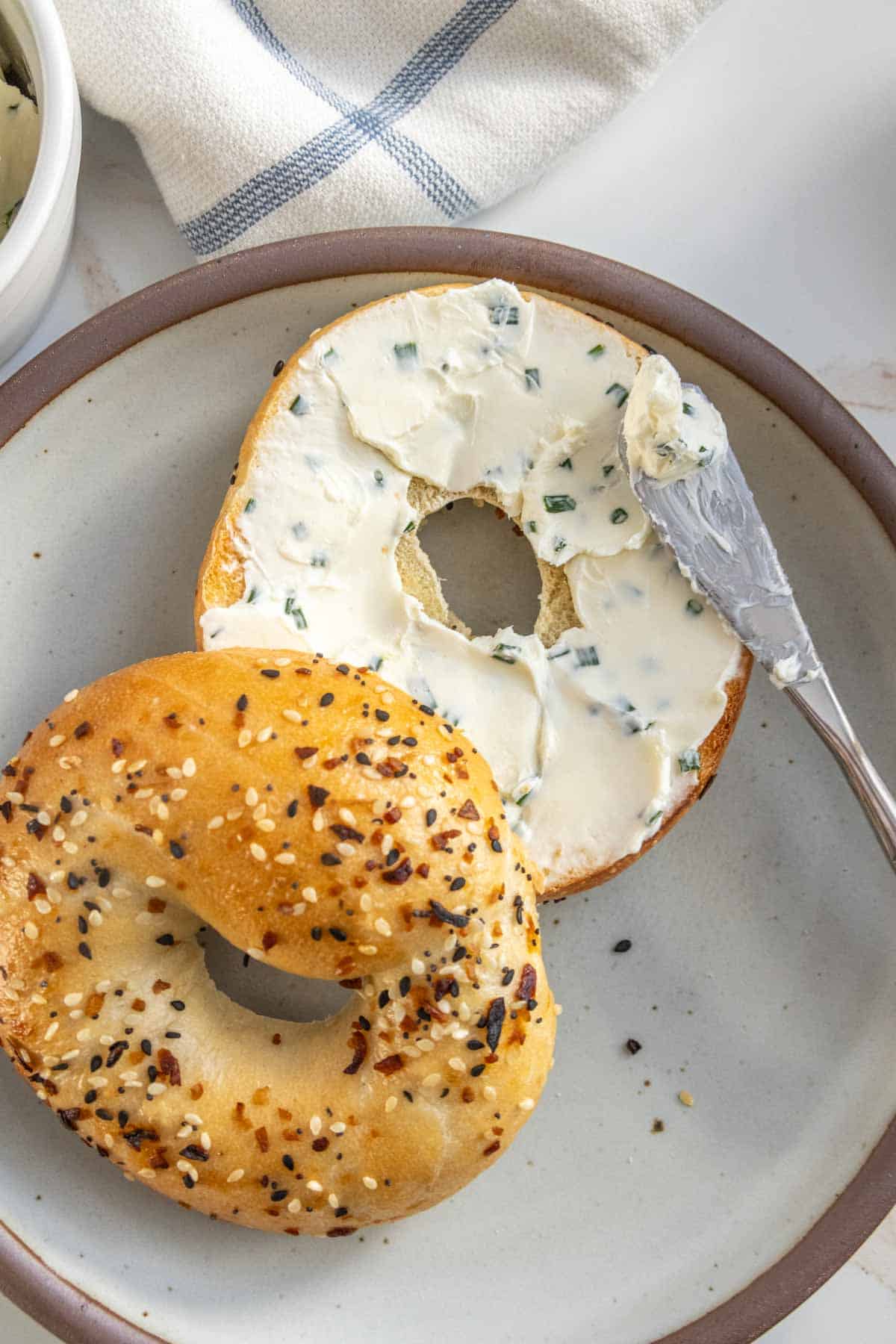 An everything bagel with cream cheese on a plate, half of it spread with cream cheese, beside a knife with cream cheese.