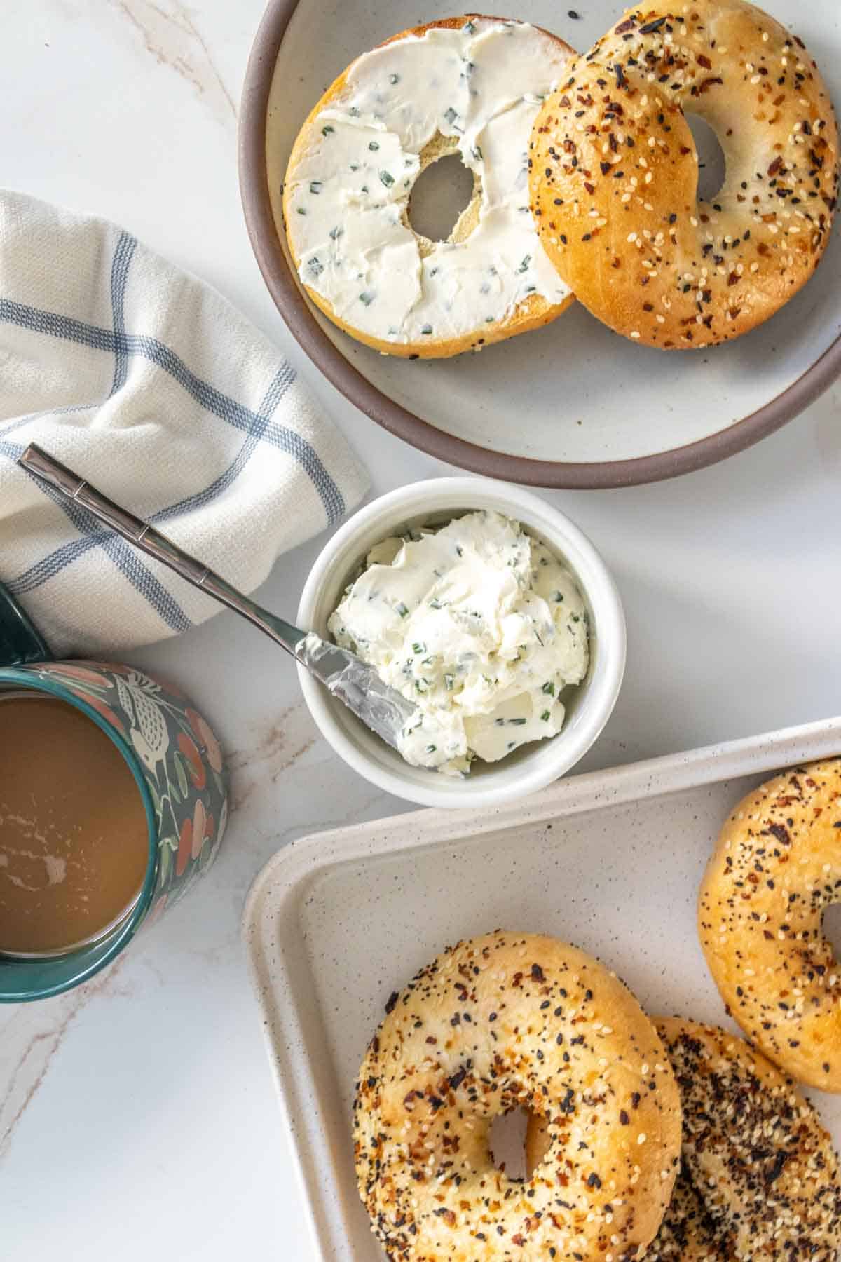 A top view of a breakfast setting featuring two plates with everything bagels, one topped with cream cheese, alongside a bowl of cream cheese and a mug of coffee.