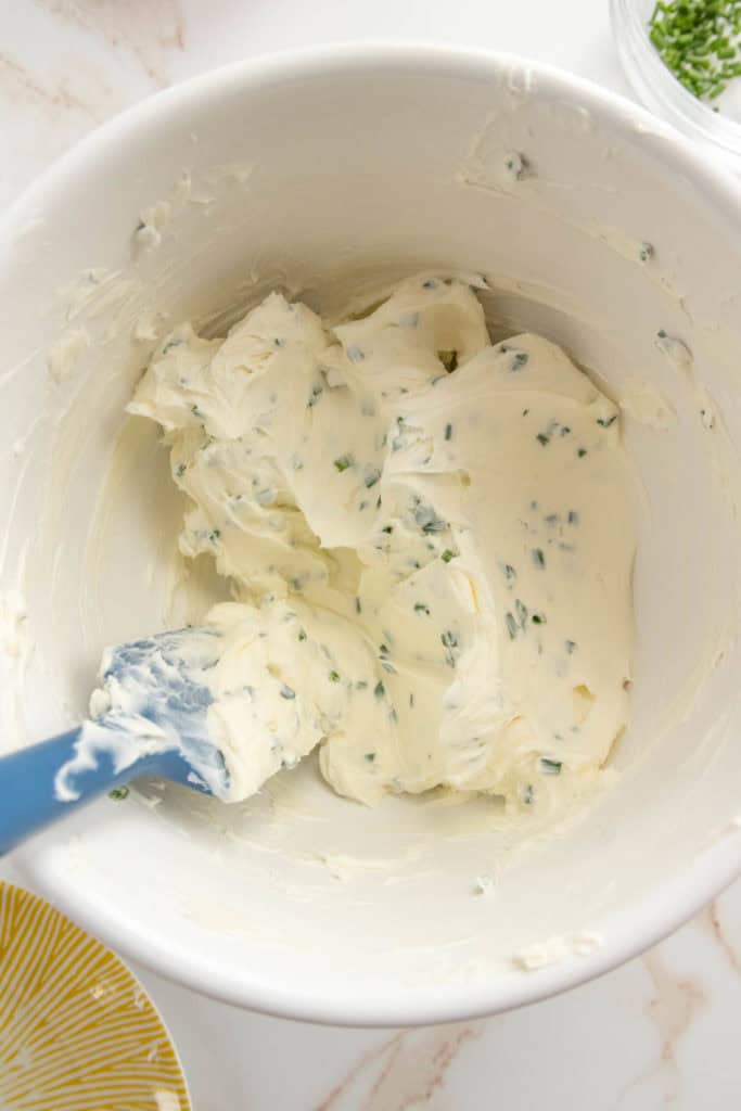 A bowl of cream cheese mixture with a blue spatula, placed on a marbled surface with a sprinkle of chopped herbs in the background.
