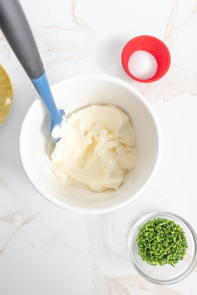 A bowl of creamy substance with a spatula, alongside a small bowl of green chopped chives and a dish of salt on a marble surface.