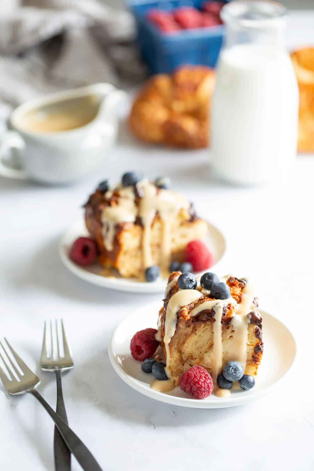 A plate with a slice of bread pudding topped with vanilla sauce and fresh blueberries and raspberries, alongside a cup of coffee and a glass of milk.