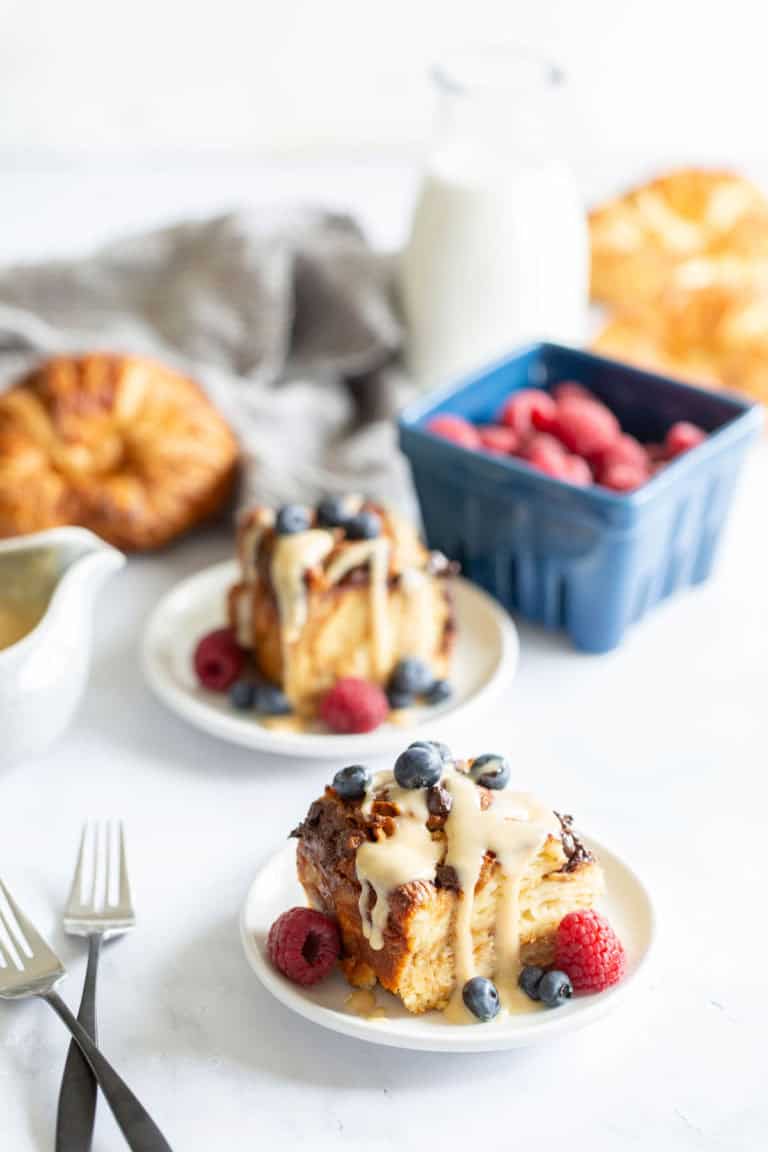A plate of croissant bread pudding topped with berries and drizzled with syrup, accompanied by a milk bottle and more berries in the background.