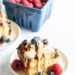 A plate of croissant bread pudding topped with cream and blueberries, served with raspberries, beside a blueberry basket and a croissant.