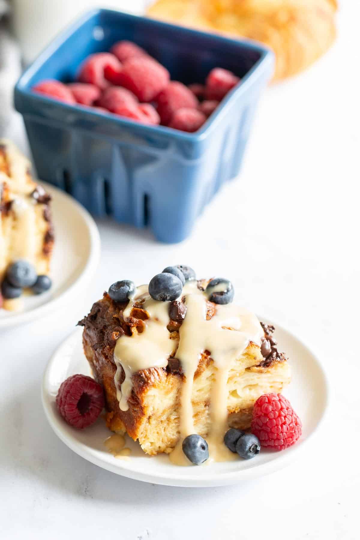 A plate of croissant bread pudding topped with cream and blueberries, served with raspberries, beside a blueberry basket and a croissant.