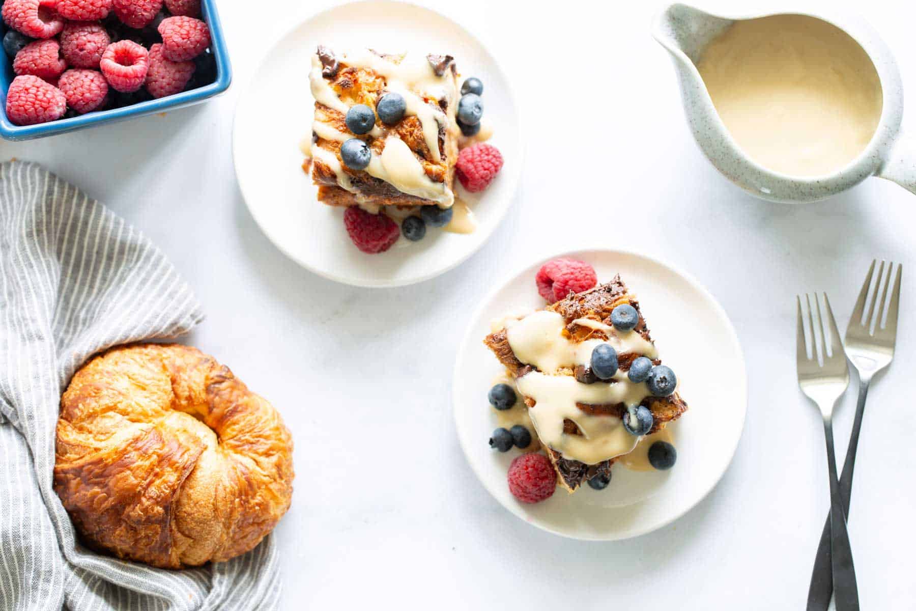 A breakfast spread with bread pudding topped with berries and cream, a croissant, and a bowl of raspberries on a white table.