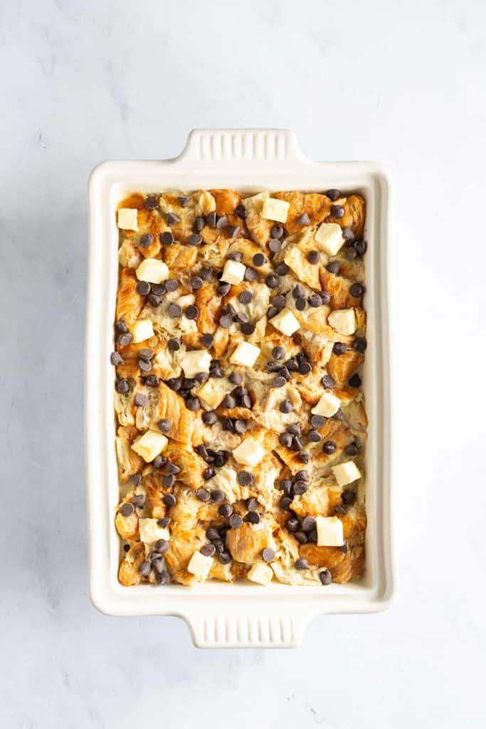 Unbaked casserole with chunks of bread, melted cheese, and chocolate chips in a white ceramic dish on a marble surface.