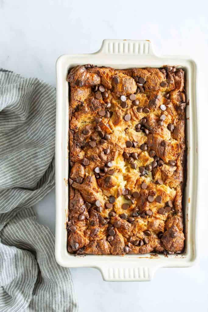 A freshly baked bread pudding with chocolate chips and nuts in a white ceramic dish on a marble countertop, next to a gray cloth.