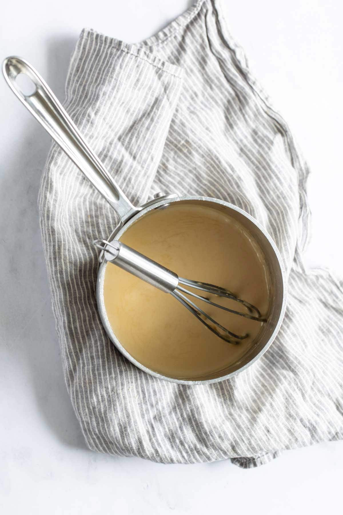 Overhead view of a saucepan containing beige sauce with a whisk, resting on a striped cloth on a marble surface.