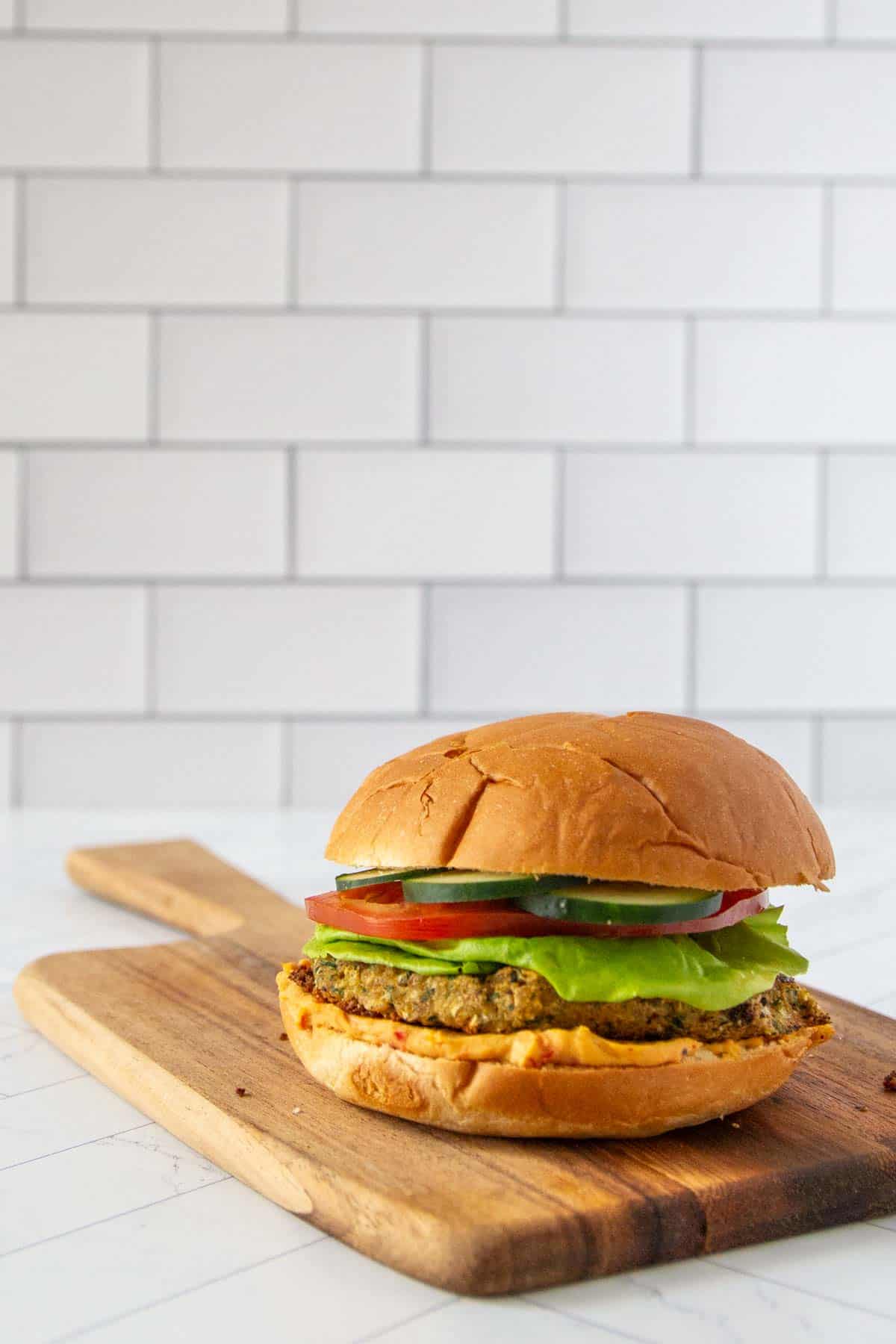 A veggie burger with lettuce, tomato, and a thick patty on a wooden cutting board against a white subway tile background.