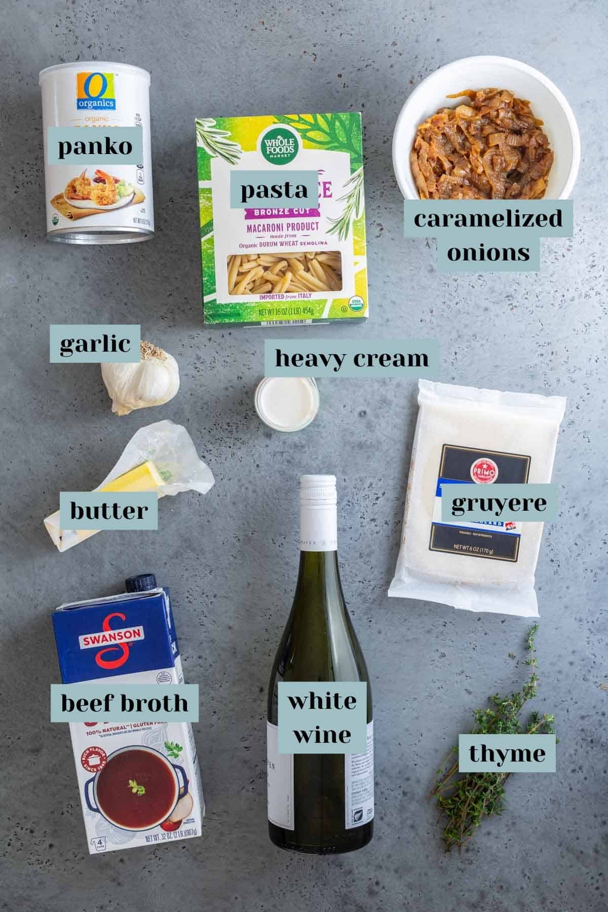 Various ingredients for cooking laid out on a gray surface, including panko, pasta, caramelized onions, garlic, heavy cream, butter, beef broth, white wine, gruyere cheese, and thyme.