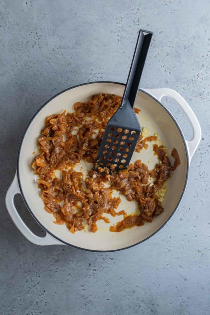 Caramelized onions in a white pan with a black spatula on a grey surface.