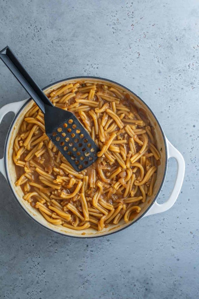 A pot of cooked pasta with sauce and mushrooms, with a black slotted spoon, on a gray surface.
