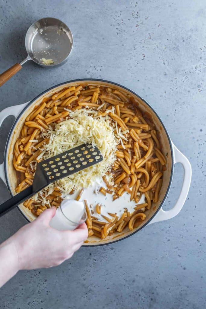A person adding shredded cheese to a pot of pasta using a cheese grater, with a small bowl of cheese and a grater handle visible on a gray surface.