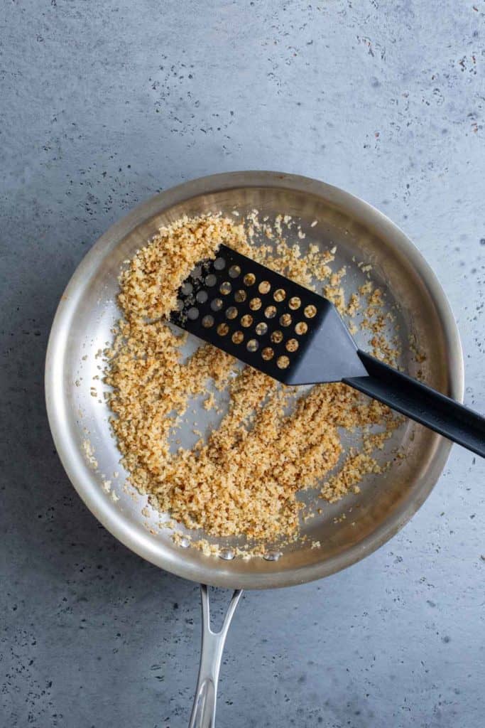 A skillet with toasted breadcrumbs and a black spatula on a speckled grey surface.
