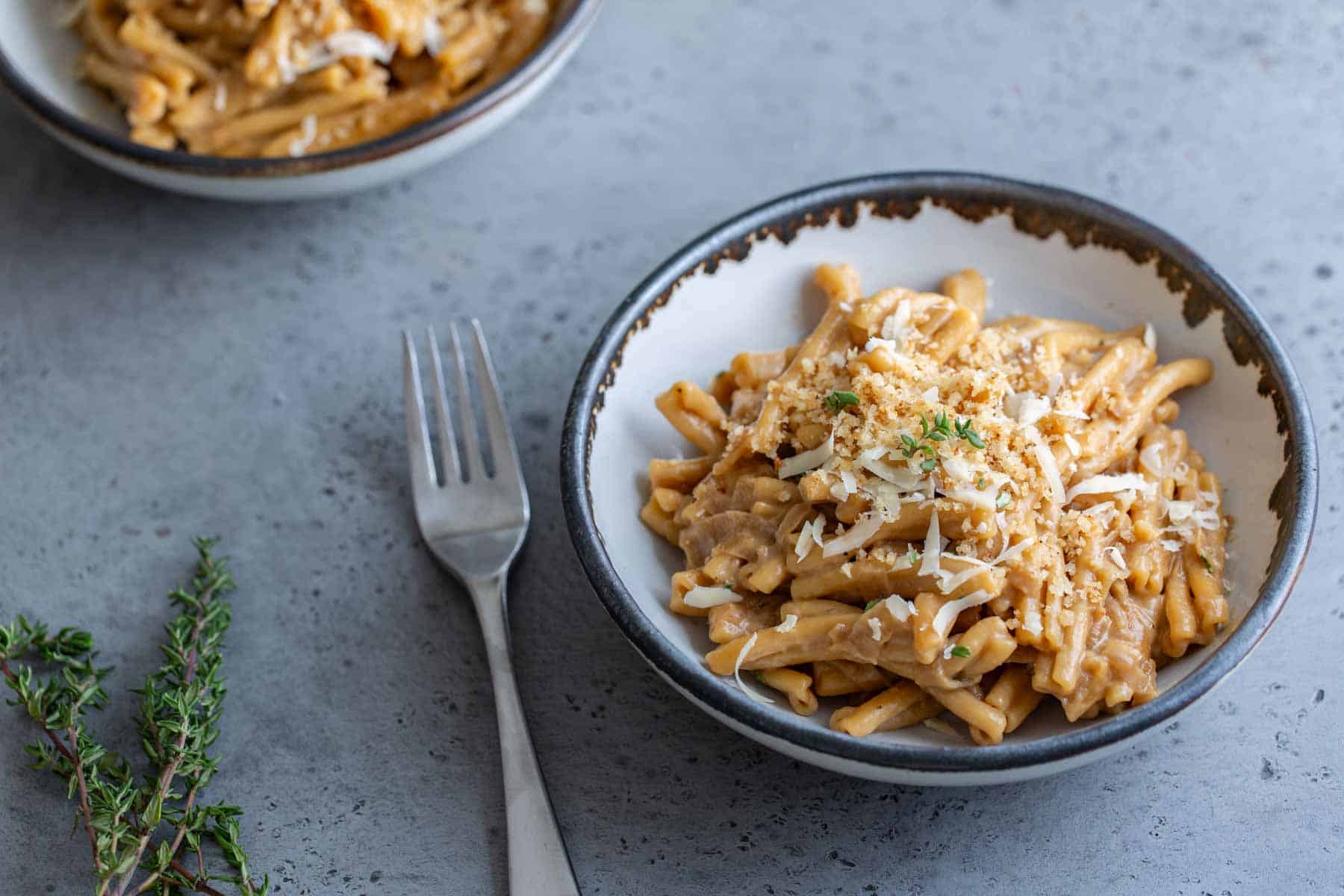 A bowl of creamy pasta topped with grated cheese and herbs, with a fork on the side and another bowl in the background.