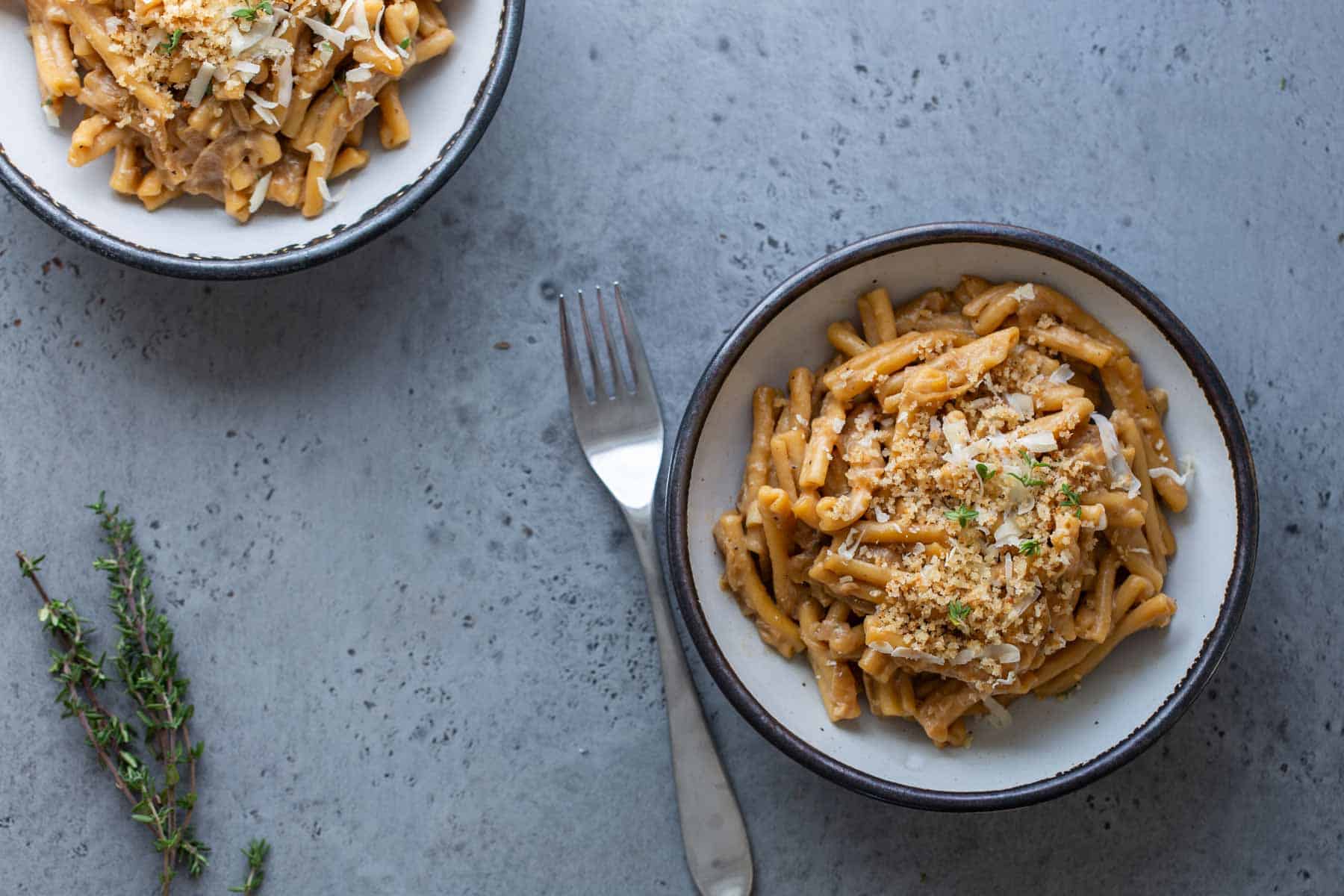 Two bowls of pasta topped with grated cheese and herbs on a textured gray surface, accompanied by a fork and sprigs of thyme.