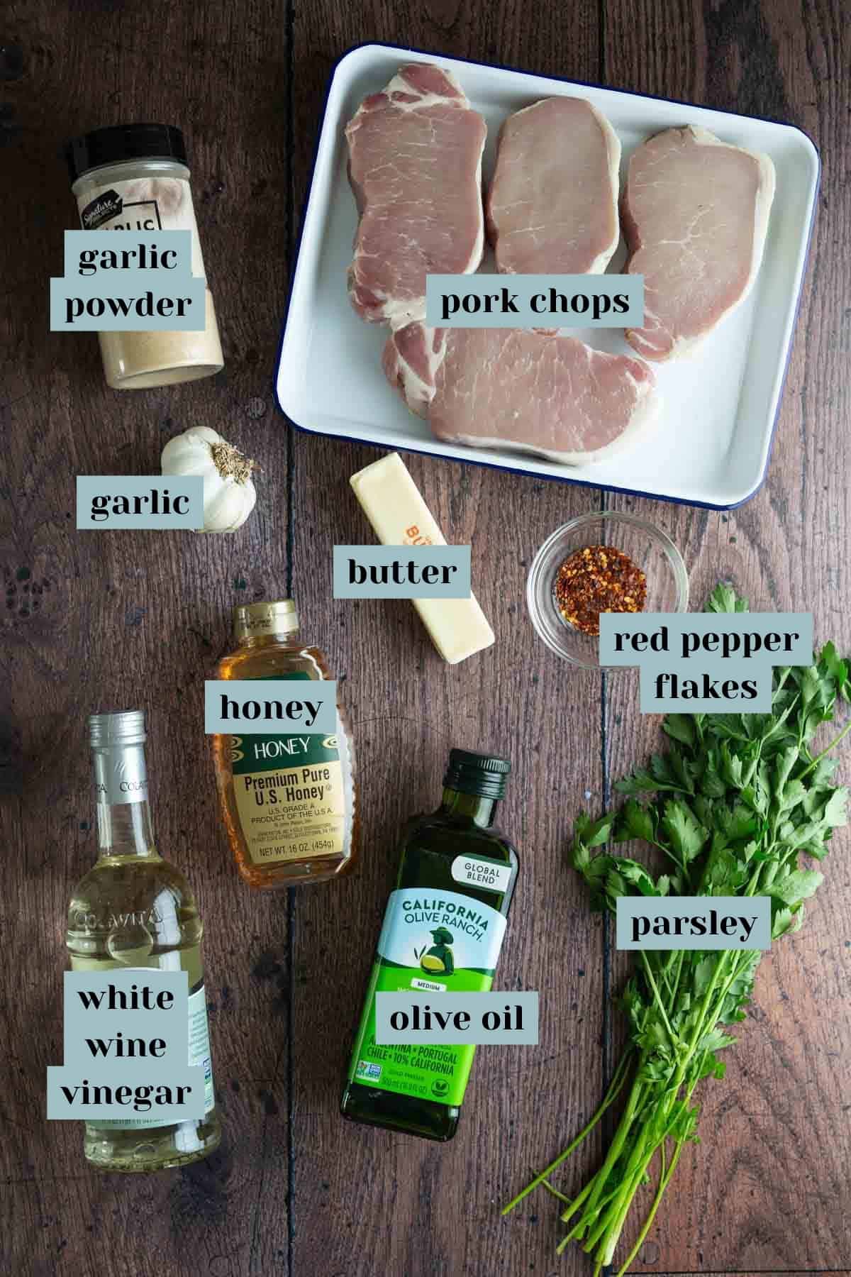 Ingredients for cooking pork chops arranged on a wooden surface, including pork chops, garlic, olive oil, honey, and spices..