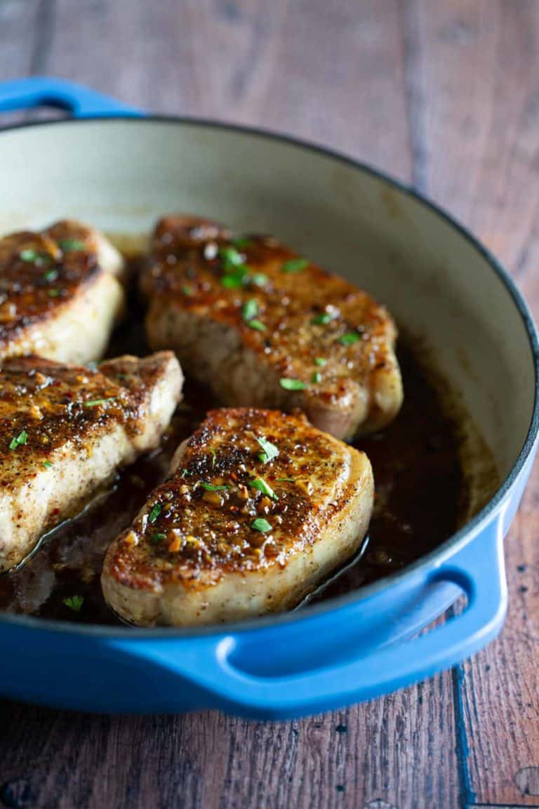 Three cooked pork chops with herbs in a blue cast iron skillet on a wooden surface.