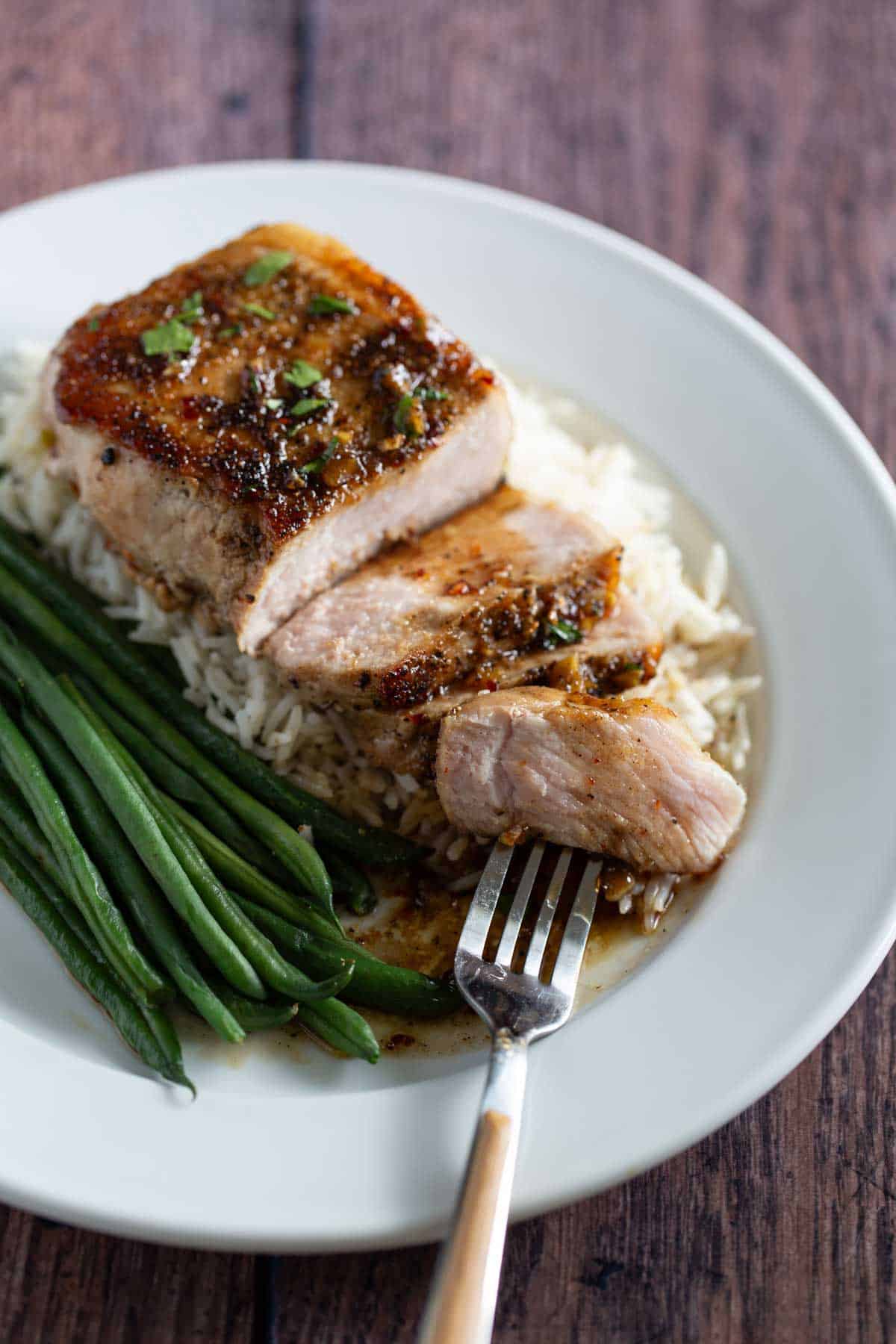 Grilled pork chop on a bed of rice with green beans, garnished with herbs, served on a white plate with a fork.
