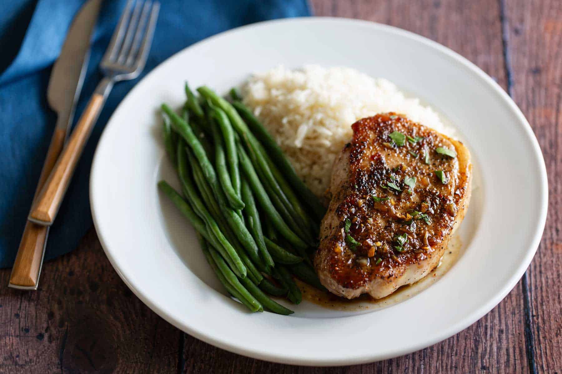 A plate with pork chop, steamed green beans, and white rice on a wooden table, accompanied by a fork and napkin.