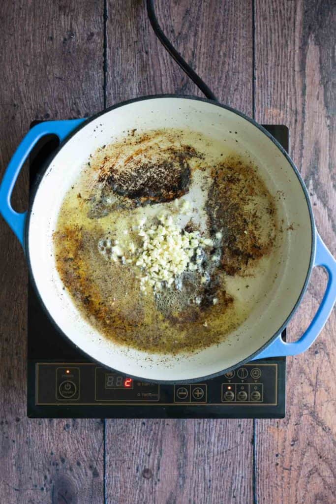 A white enameled skillet on an induction stove, with burnt remnants and chopped garlic in melted butter.