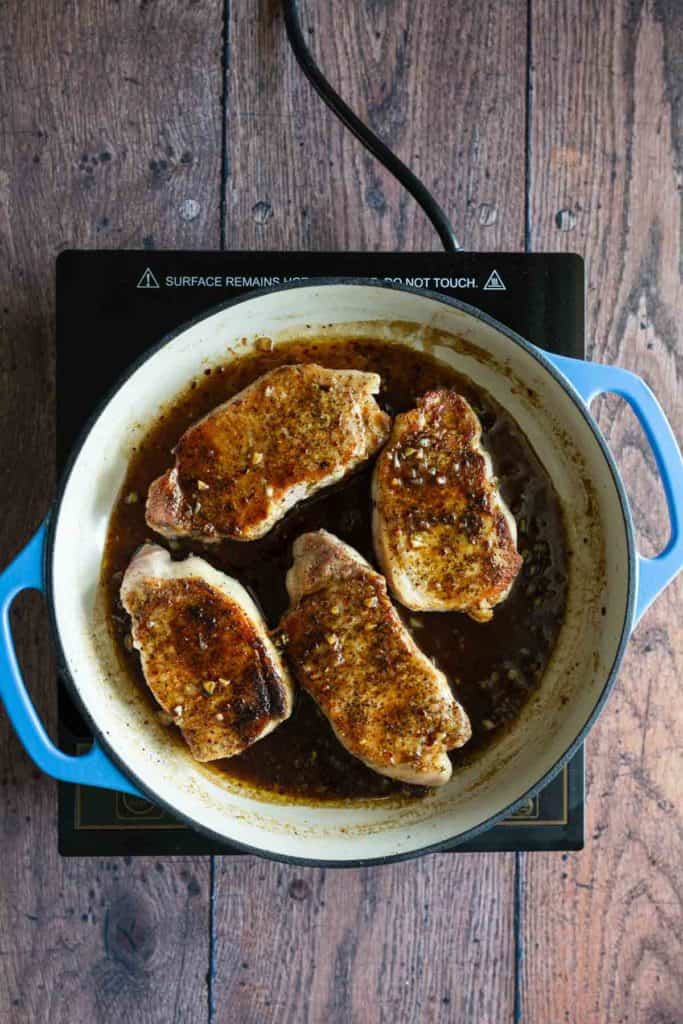Four pork chops cooking in a white enameled cast iron pot on an induction cooktop, viewed from above.