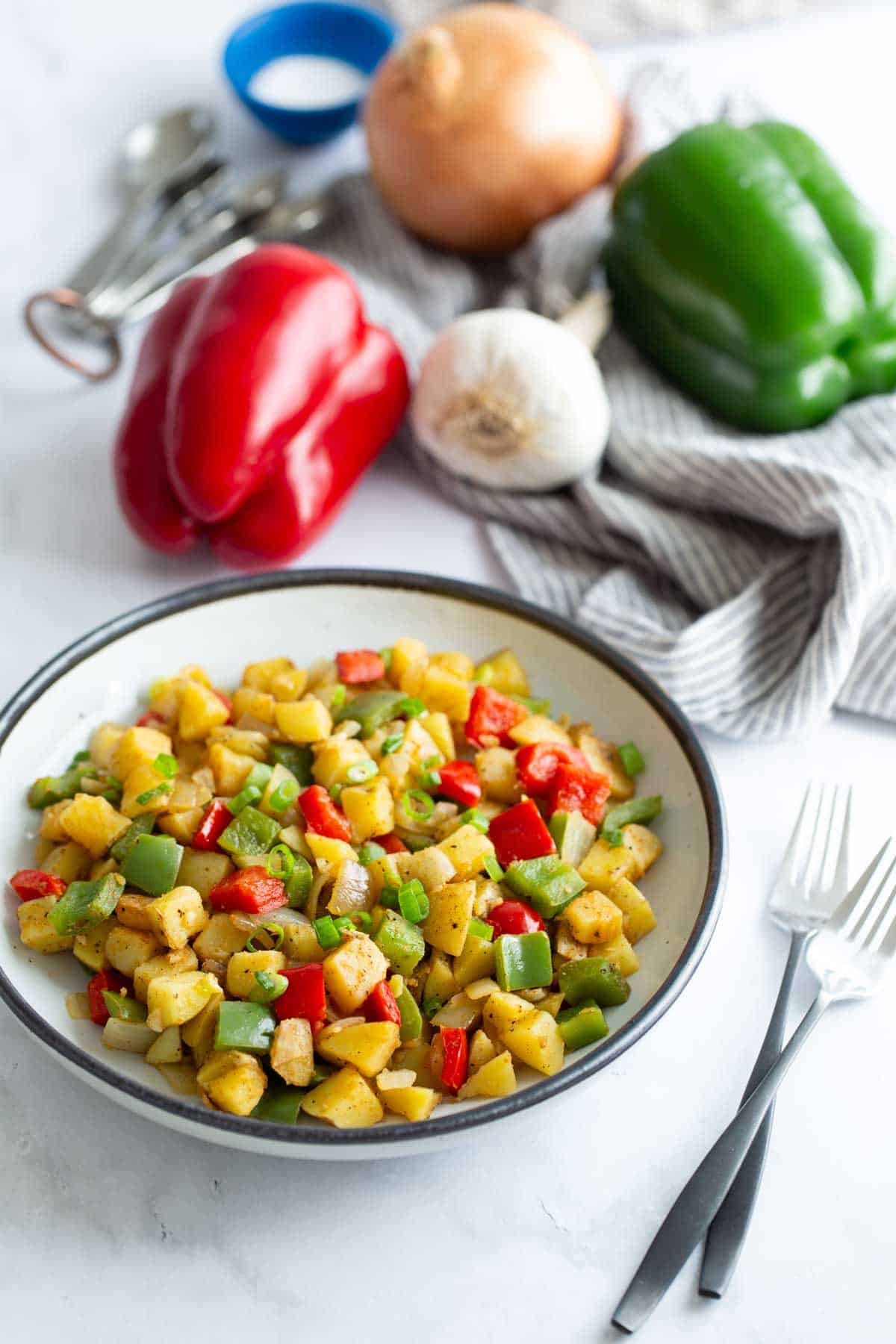 A bowl of sautéed diced potatoes with red and green bell peppers, with whole vegetables and measuring cups in the background.