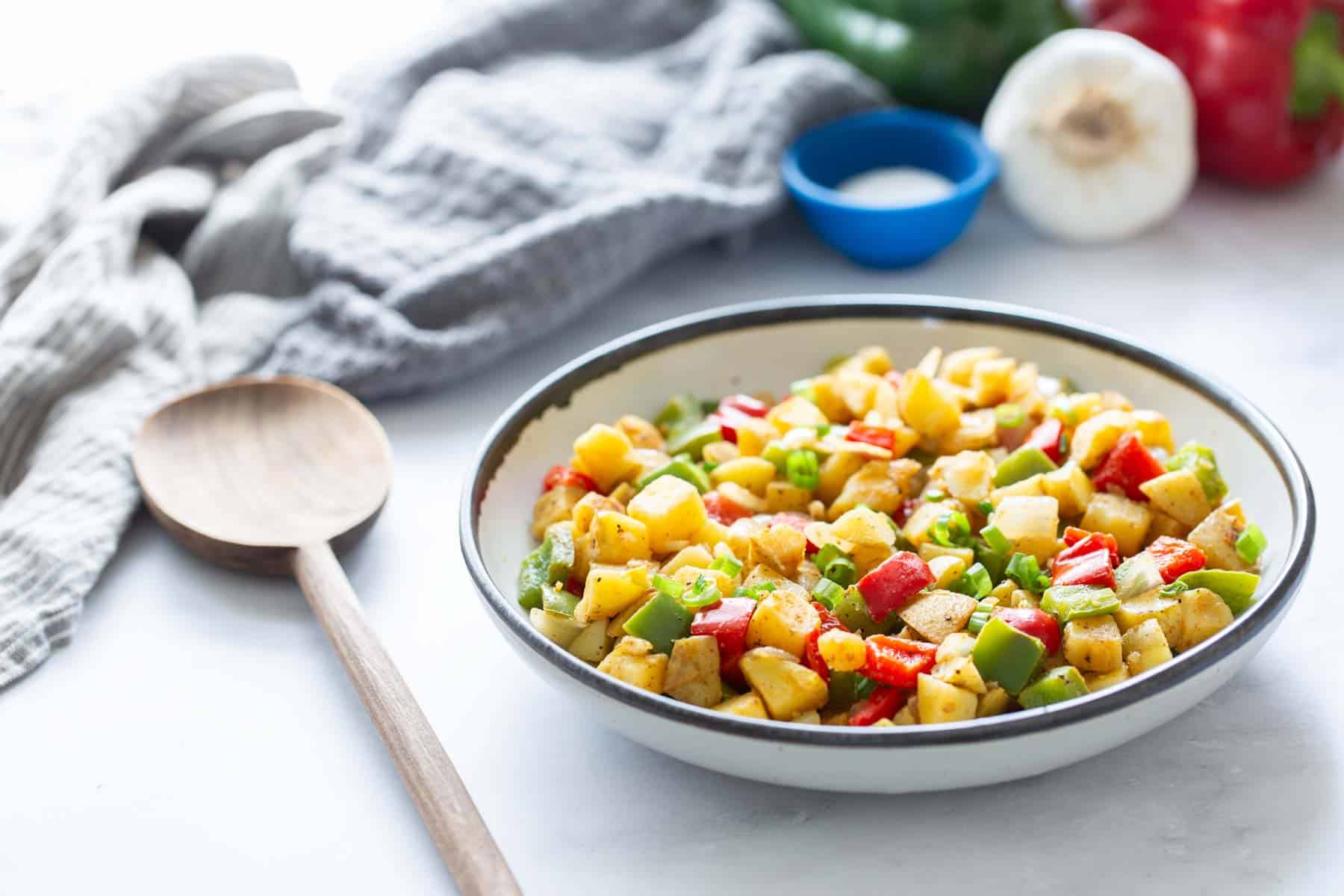 A colorful sautéed vegetable dish in a pan, with fresh ingredients on the side and a wooden spoon.