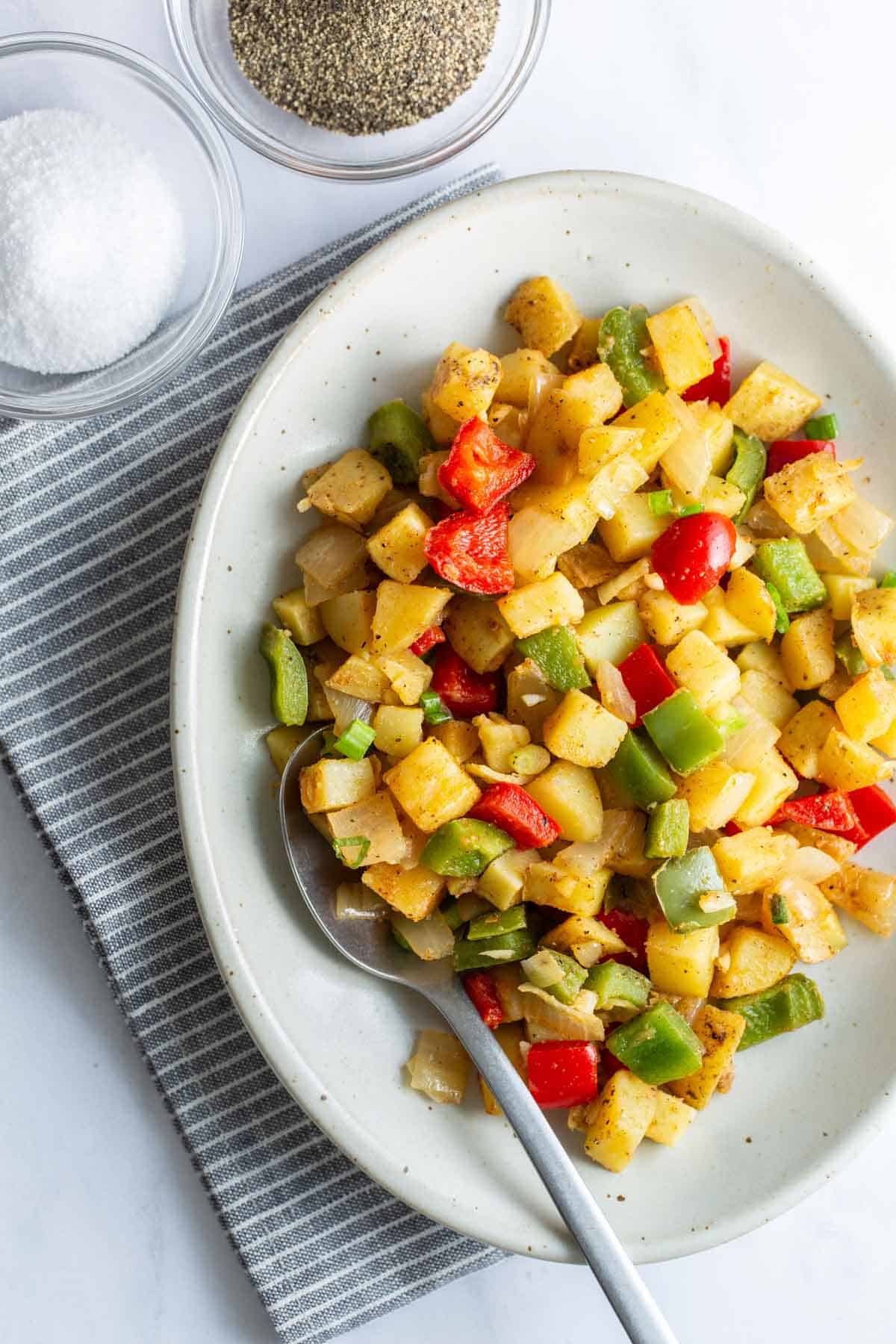 Potatoes with peppers served on a plate beside a spoon, with salt and pepper to the side.