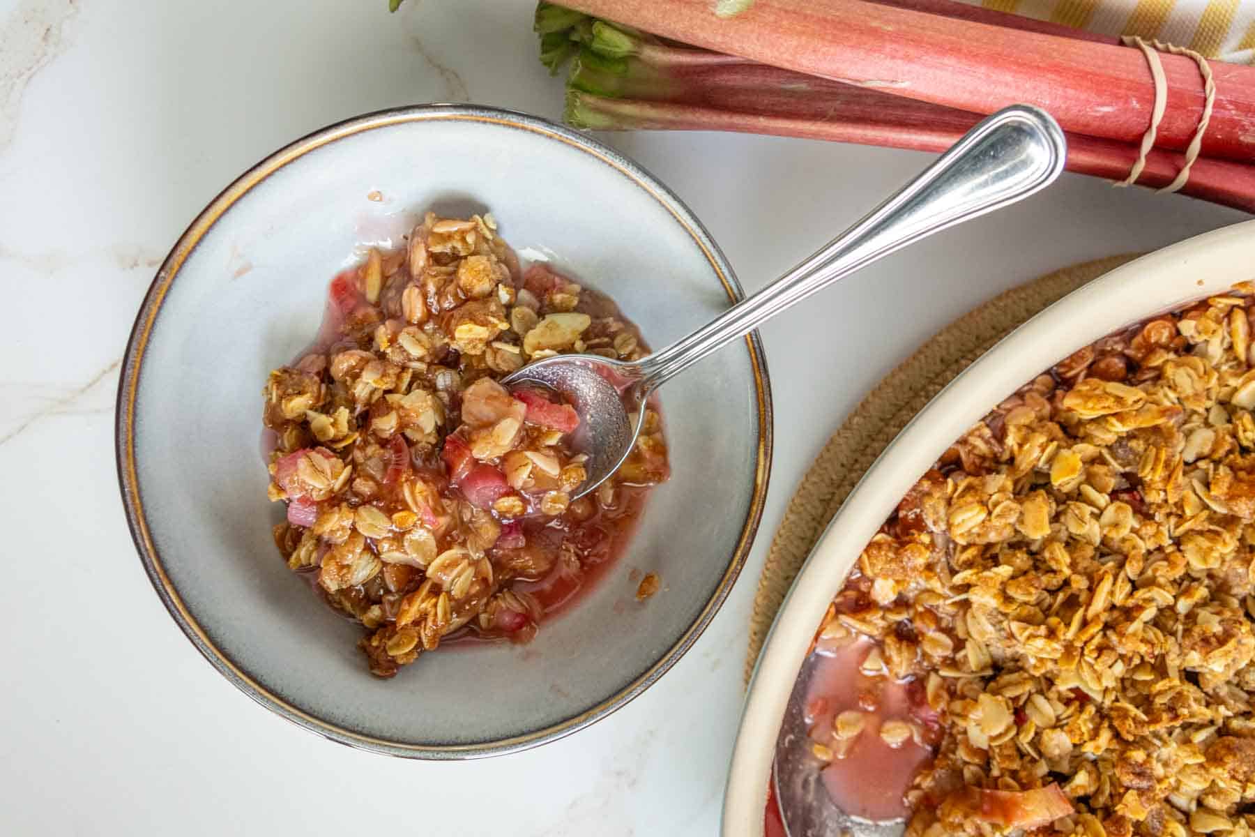 A bowl of rhubarb crumble with a spoon, beside a larger dish of the same dessert and fresh rhubarb stalks on a marble surface.