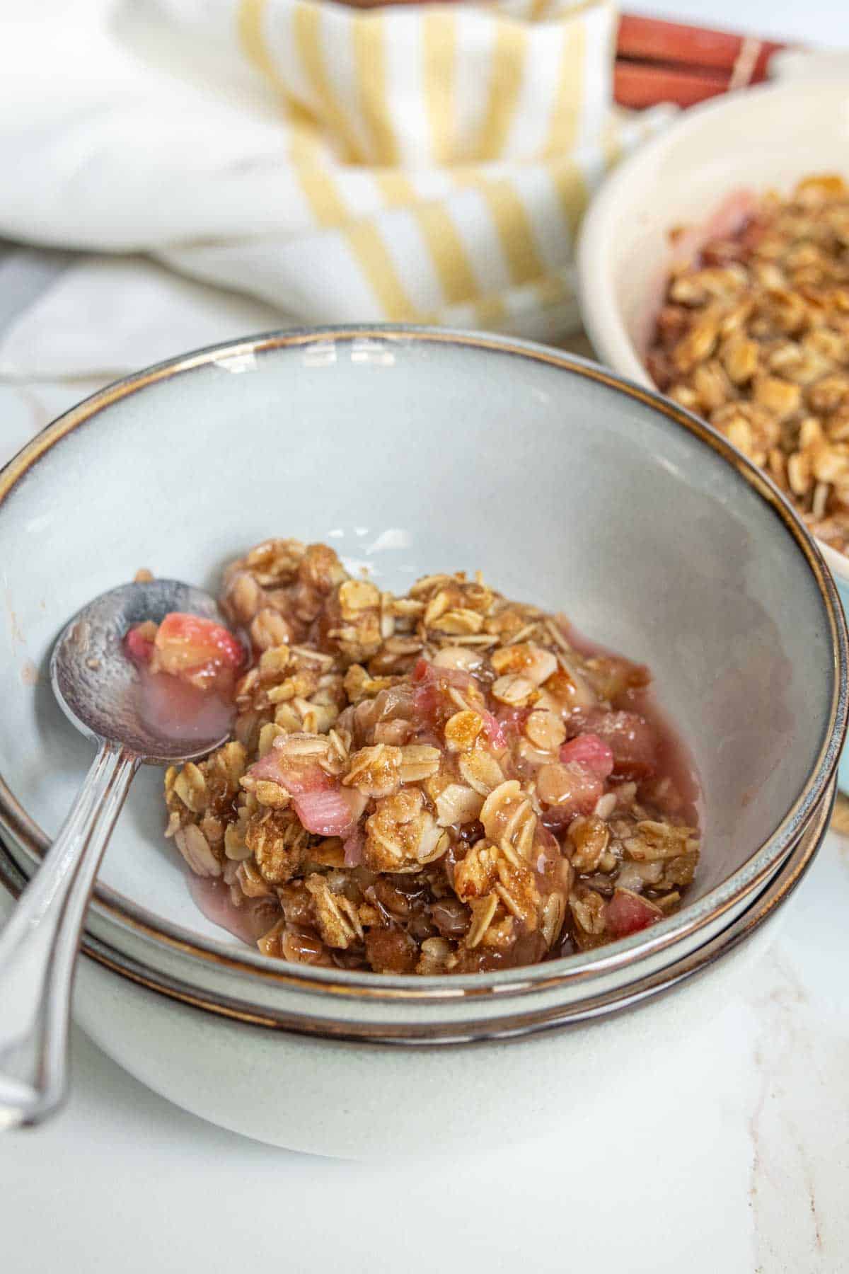 A bowl of rhubarb crisp, accompanied by a spoon, on a white surface with a striped napkin in the background.