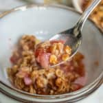 A spoonful of rhubarb crumble being lifted from a bowl, showcasing the textured topping and chunky fruit filling.