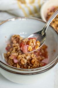 A spoonful of rhubarb crumble being lifted from a bowl, showcasing the textured topping and chunky fruit filling.