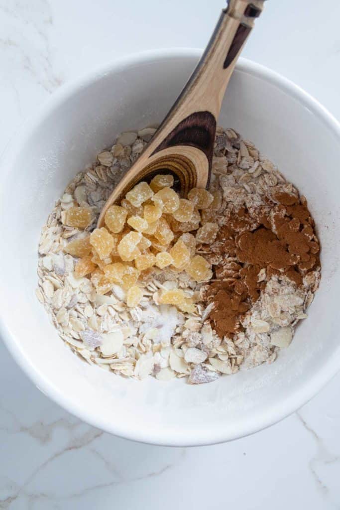 A white bowl containing oats, sliced almonds, chopped ginger, and cinnamon, with a wooden spoon on a marble surface.