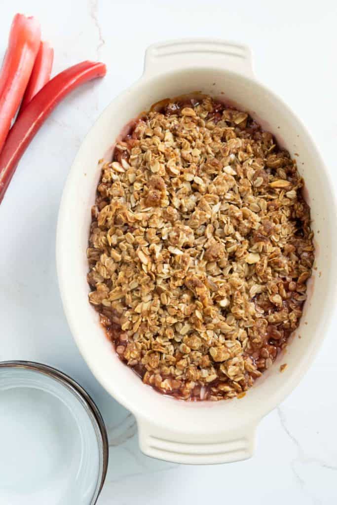 Baked rhubarb crisp in a white oval dish, topped with a golden-brown oat crust, served on a marble countertop with fresh red rhubarb stalks and a glass of water beside it.