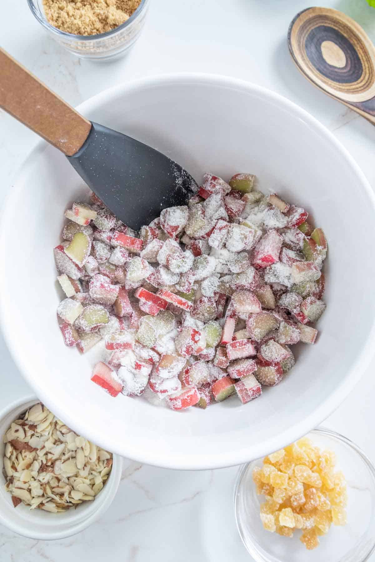 A bowl of chopped rhubarb mixed with sugar, with a spatula, surrounded by ingredients like almonds and crystallized ginger on a marble surface.