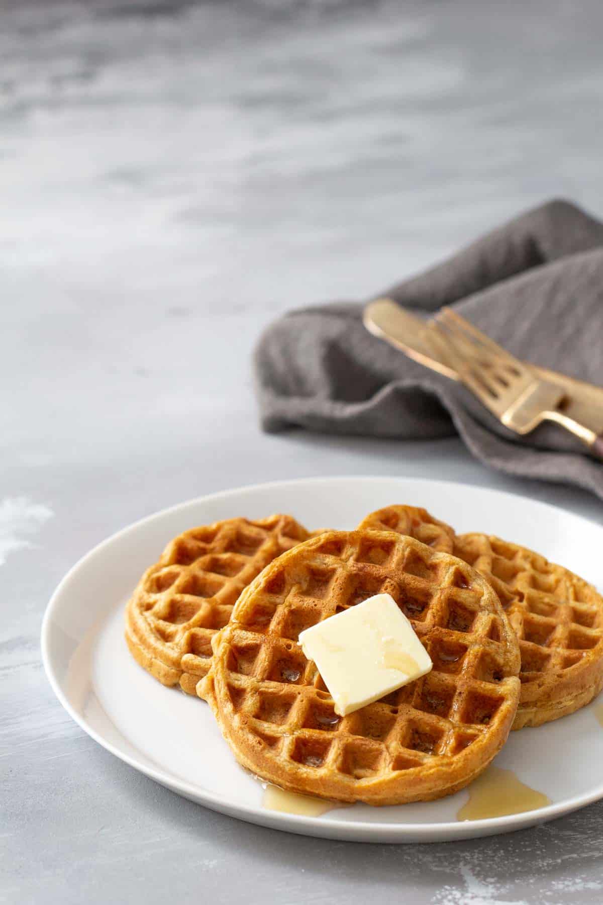 A plate with freshly baked waffles topped with a pat of butter, accompanied by a fork and knife on a napkin.